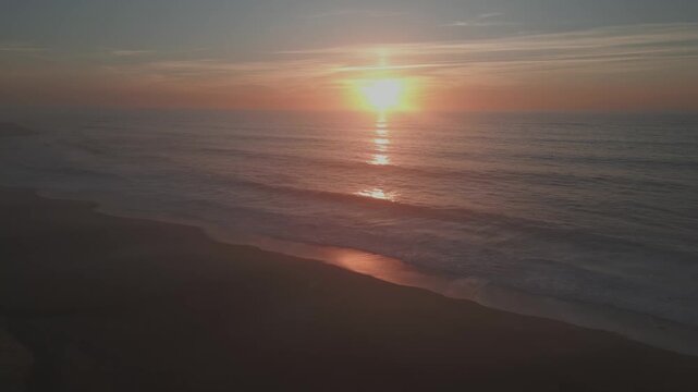 Cinematic slow motion aerial view of a vibrant sunset over the Atlantic Ocean at Praia do Salgado in Nazar&eacute;, Portugal. Calm waves wash over the dark sand in the warm evening light