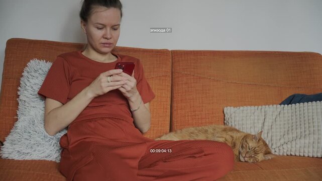 Young woman scrolling smartphone on sofa with ginger cat, cozy home lifestyle, casual browsing, medium shot