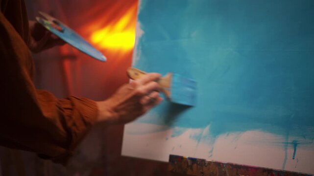 Artist Skillfully Applying Multiple Layers Of Vibrant Blue Color On Textured Canvas In Dim Lighting