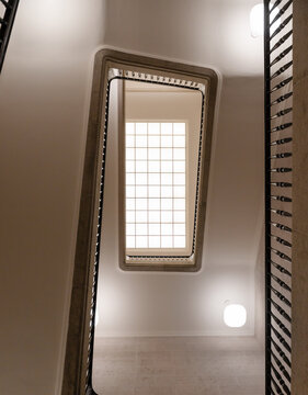 Spiral rectangular staircase - low angle view. An upward perspective of a modern minimalist staircase with a central skylight, wrought iron railings, and warm ambient lighting in an elegant interior.