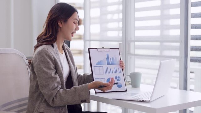 Adult Asian businesswoman presenting data charts during virtual online meeting from modern home office workspace