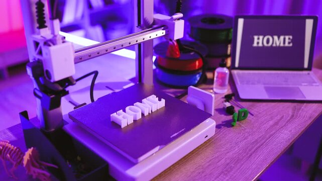 A 3D printer is actively creating the word HOME in white plastic, with spools of filament and a laptop displaying the same word in the background