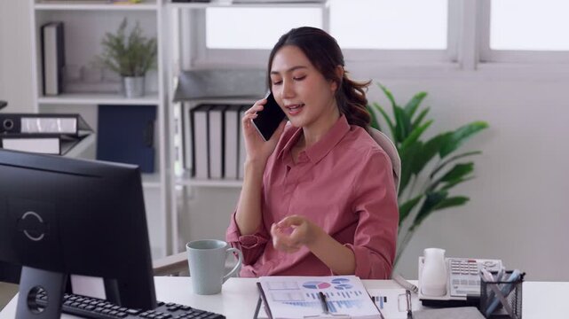 Asian business professional young adult female entrepreneur discussing business on mobile phone communicating effectively working remotely at modern home office workstation