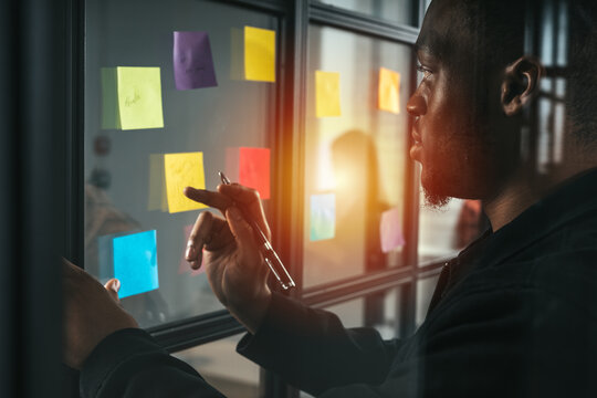 Man working on glass board with colorful sticky notes in an office during sunset while holding a pen