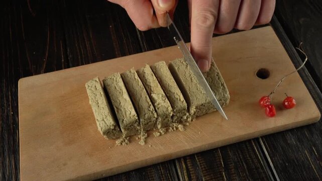 Hands skillfully cut a block of halva into identical pieces on a wooden cutting board, placing viburnum berries next to it, emphasizing the culinary taste of the preparation in the kitchen