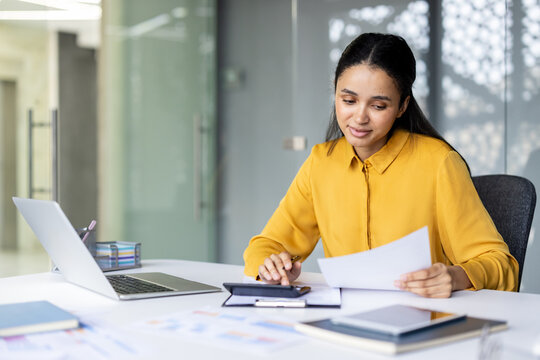 Young professional woman or accountant sitting at an office desk, analyzing financial documents and using a calculator while working with a laptop and other paperwork, managing business finances