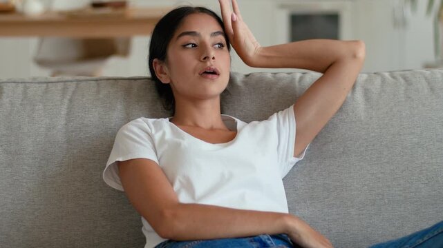 Sad upset Indian Arabian woman at home waving hands feeling overheated frustrated female suffering high temperature inside living room broken air conditioner stressed sweaty sofa summer heat indoors