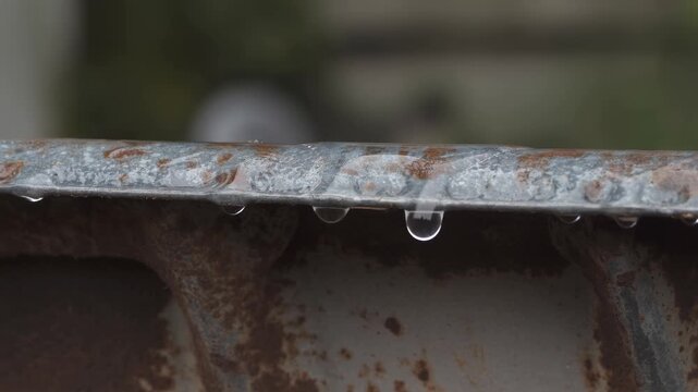 Close-up of water drops dripping from a rusty metal edge. Rainwater falling from old corroded iron surface. Weather and decay concept