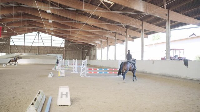 Skilled female jockey riding a beautiful brown horse, jumping over an obstacle in an indoor arena
