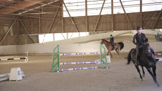 Two professional riders training horses for show jumping in a large, sandy indoor equestrian arena