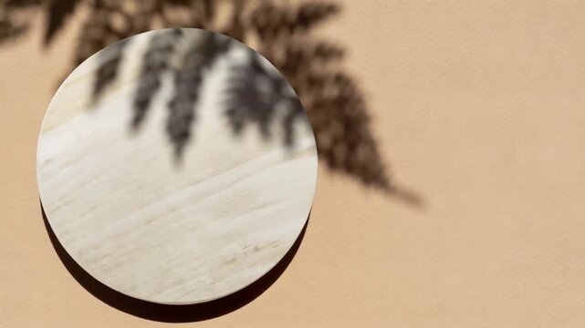 An empty white round rotating podium for product demonstration with the shadow of a palm leaf on a beige background.