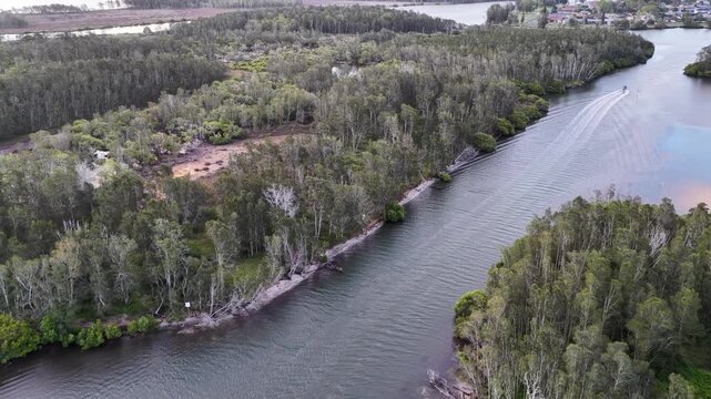 Aerial drone video over Pipers Bay in Forster, New South Wales, Australia showing a boat travelling through the channel with the sun setting behind a cloudy sky.