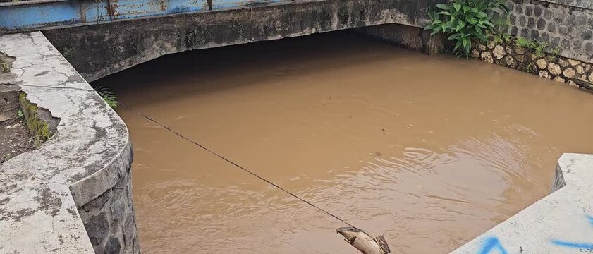 Murky, brown floodwater flows quickly through a concrete-lined canal under a bridge structure. The muddy river indicates heavy rainfall or flooding upstream.