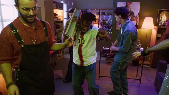 Full length shot of cheerful young adult African American man dancing to music with boombox on shoulder while having fun with friends at home party
