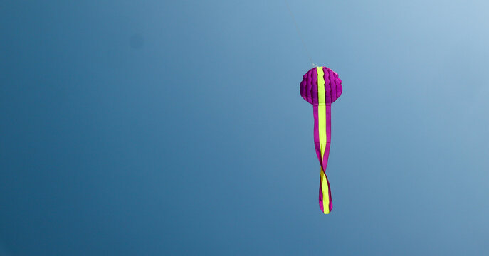 A long purple kite with yellow stripes soars high against a clear blue sky, its long pink and yellow tail ribbons twisting gracefully in the wind.