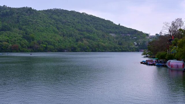 Time-lapse view of Naukuchiatal Lake, Naukuchiatal, highlighting rippling waters with a lakeside activity zone featuring paddle boats and a transparent water roller, set against forested hills.
