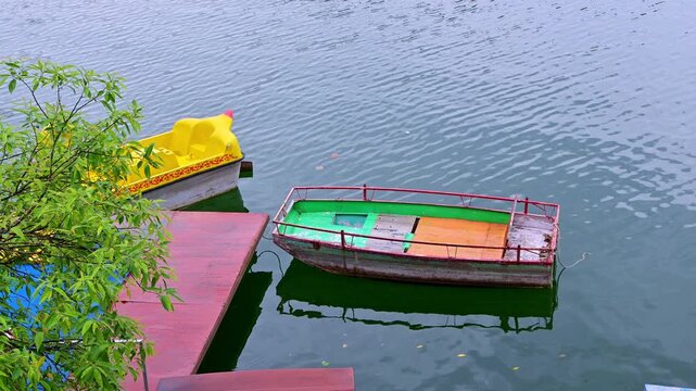 A calm lakeside scene in Naukuchiatal India shows a weathered multicolored rowboat tied beside a red wooden dock, with a bright yellow pedal boat partially hidden by leafy branches, floating on water.