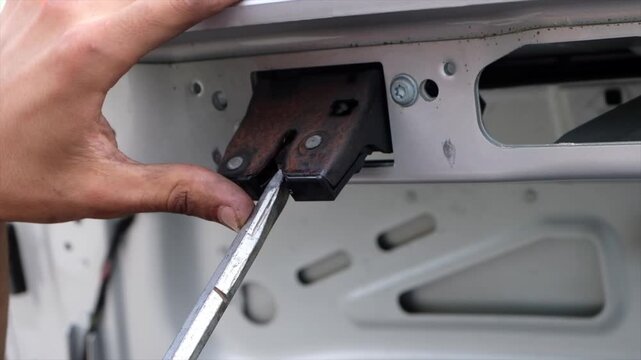 Mechanic hand testing rusty car trunk latch using metal tool for repair maintenance service process.