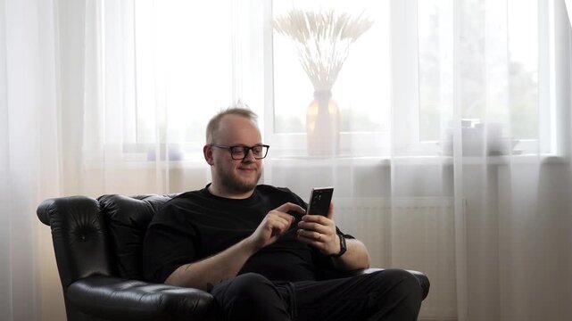 Happy man with glasses using smartphone at home, smiling while reading good news in cozy living room
