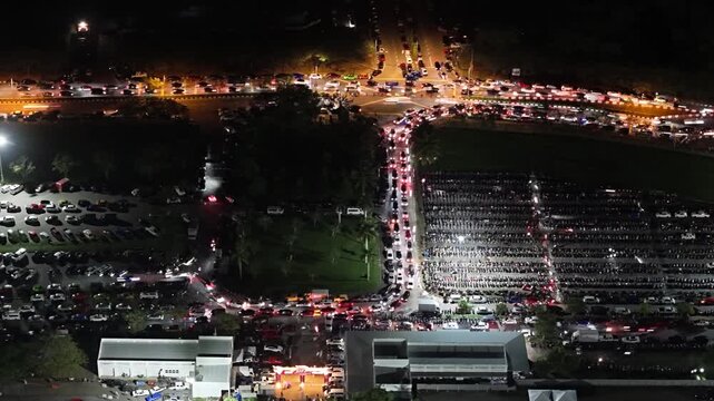 Aerial night view of vehicle light trails and parked cars at a congested intersection and parking lot at Batu Kawan Stadium