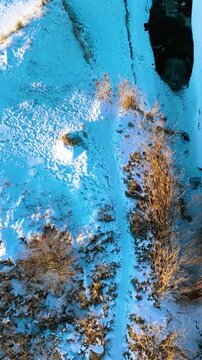 Vertical aerial shot showing a dark frozen river cutting through a snowcovered landscape with dry reeds and animal tracks. Useful for nature documentaries and winter climate themes.