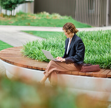 Portrait of a young white businesswoman woman using a laptop computer and sitting on a bench in a coty park, surrounded by modern corporate office buildings