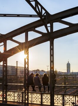 Three youths sitting on the balustrade of the Hackerbr&uuml;cke bridge over the railway tracks looking into the distance, back light, Munich, Upper Bavaria, Bavaria, Germany