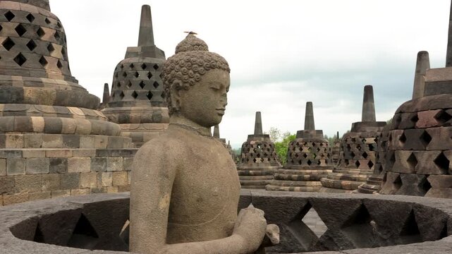 Horizontal panning of stone Buddha statue seated inside a perforated stupa at Borobudur temple in Java, Indonesia. Ancient Buddhist monument with multiple latticed stupas and spires