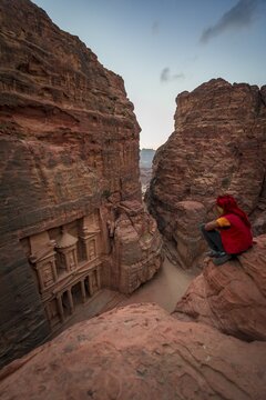 Bedouin sits at the edge of a rock and looks from above into the gorge Siq, Pharaoh's treasure house carved into the rock, facade of the treasure house Al-Khazneh, Khazne Faraun, mausoleum, Nabataean city Petra, near Wadi Musa, Jordan