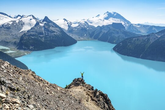 Young man standing on a rock, stretching his arms in the air, view of mountains and glacier with turquoise blue lake Garibaldi Lake, peaks Panorama Ridge, Guard Mountain and Deception Peak, Garibaldi Provincial Park, British Columbia, Canada