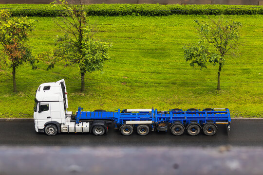 White semi truck with blue container chassis moves past green roadside vegetation, illustrating freight transport and modern logistics operations. Scene highlights structure of container transport equ