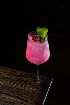 Colorful pink cocktail with mint leaves and flower on wooden table in a dim setting during evening hours
