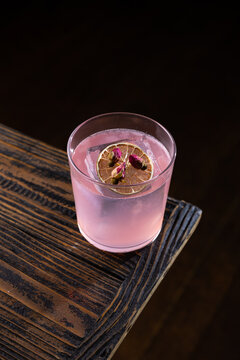 Bright pink drink on wooden table with dried lemon slice and flower garnish in dark setting