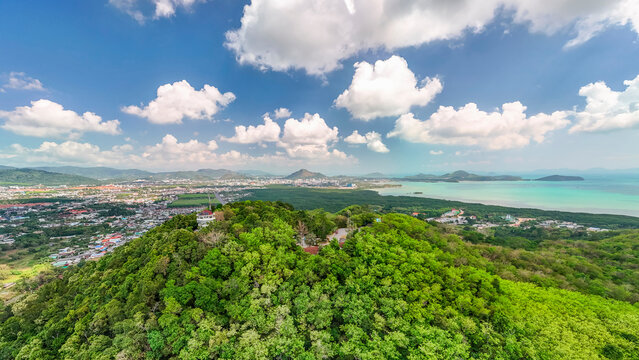 From Kaokard Viewpoint Tower, a stunning vista unfolds over Phuket city, a tranquil bay, and distant mountains. Lush green forest envelops the tower under a bright blue sky filled with white clouds.