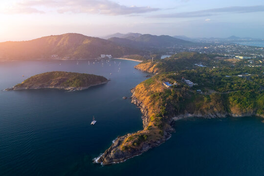 Aerial view of Phuket's dramatic coast with an offshore island, yachts on calm waters, and a vibrant sunset