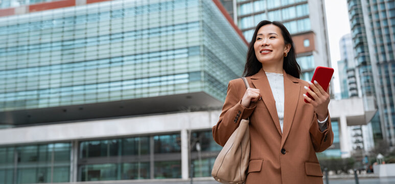 Asian businesswoman using smartphone on city street, professional career woman checking online banking and finance apps, walking at business office buildings in urban downtown. Web banner, copy space