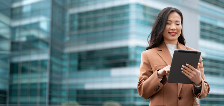 Asian entrepreneur woman CEO holding touchscreen gadget device. Smiling businesswoman using digital tablet outdoors at office building, corporate technology, mobile work. Web banner, copy space