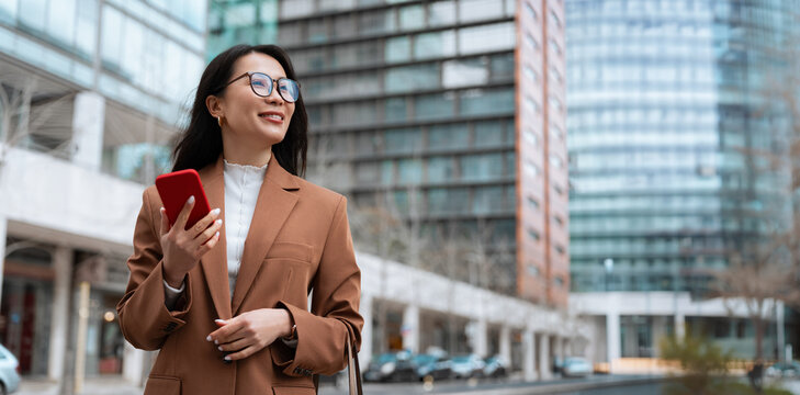 Smiling businesswoman in brown blazer holding cell phone smartphone in modern city street at office buildings, looking away. Asian busy woman using phone. Urban lifestyle. Web banner, copy space