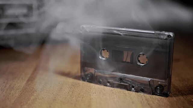 A clear audio cassette tape sits on a wooden desk as thick white smoke billows around it. Concept of retro technology overheating, musical nostalgia, and analog destruction.