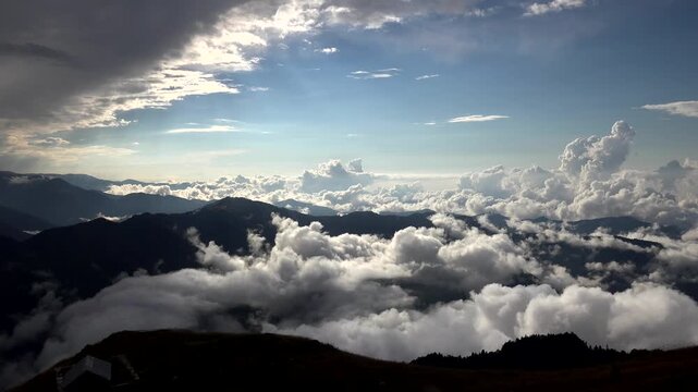 Mountain timelapse above clouds with cabin on alpine ridge at sunrise under dramatic sky. Misty peaks and highland shelter emerge through morning fog beneath glowing dawn horizon.