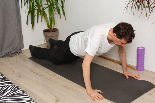 A man, 53 years old, lies on a yoga mat at home, performing a stretching exercise. He focuses on his fitness routine in a simple indoor space with plants around