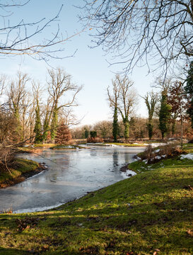 Frozen lake and winter landscape in Babelsberg Park Potsdam. Scenic view of a partially frozen lake surrounded by winter trees and green grass at Park Babelsberg. Feeling of winter changing to spring.