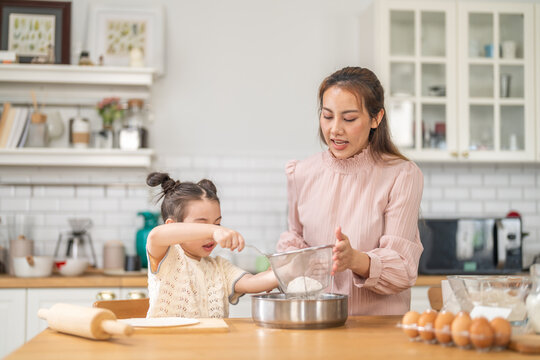 Asian happy mother and little daughter sifting flour with sieve into bowl in kitchen, Family home cooking, baking bakery, childhood learning, motherhood bonding, domestic lifestyle food concept