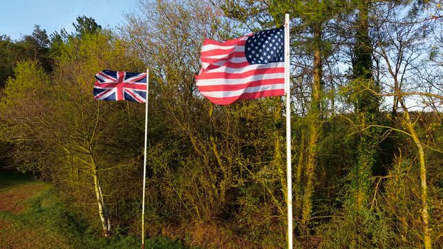 United States and United Kingdom flags waving on flagpoles at military memorial symbolizing remembrance and allied unity. Wind moving stars stripes and Union Jack under warm sunlight among trees