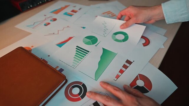 A person is sorting through various printed graphs and charts on a desk. The workspace is filled with papers showcasing different types of data visualizations and a notebook