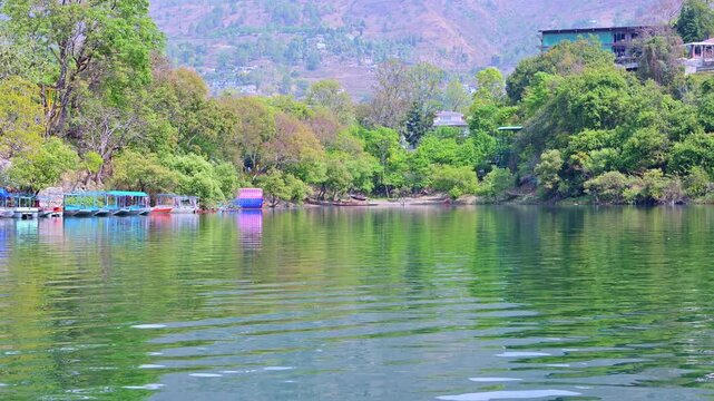 A tranquil corner of Naukuchiatal Lake, India, reflects lush green trees and distant hills, with a line of covered boats and a colorful water zorb resting quietly along shaded shoreline.