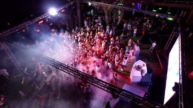 Drone aerial view captures outdoor night disco party crowd dancing around DJ stage. Energetic open air scene shows light beams spotlights pulsing wild music vibes useful lifestyle context.