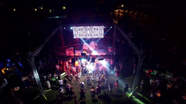 Drone aerial view captures outdoor night disco party crowd dancing around DJ stage. Energetic open air scene shows light beams spotlights pulsing wild music vibes useful lifestyle context.