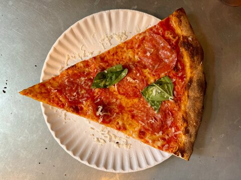 Top-down close-up of triangular slice of pizza with spicy salami and basil leaf on metal counter in pizzeria in New York City