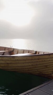 Vertical video: Sun diffusing through mist, wooden rowboat slowly bobbing at dock, water reflecting